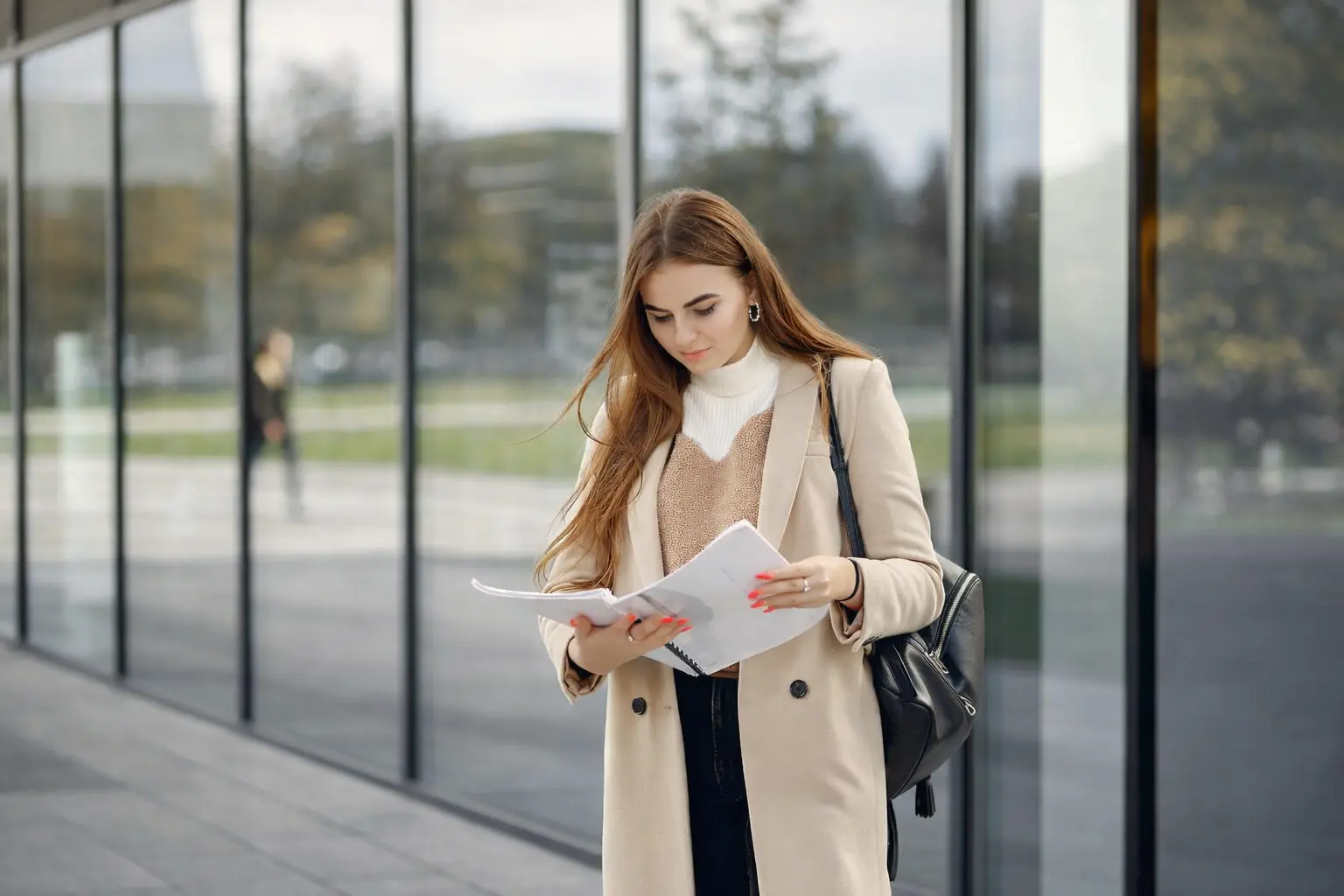A realistic editorial photograph of a professional woman in a tan coat reviewing financial documents outside a modern glass building, symbolizing the focus required for financial stability
