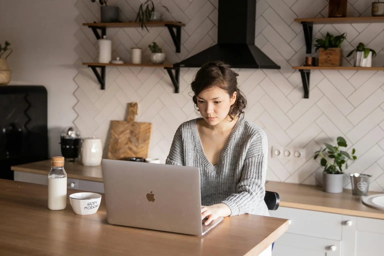A pensive person at a wooden kitchen table reviewing household bills while a high-growth city with glowing financial charts is visible through the window.