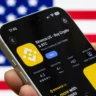 Smartphone displaying the Binance.US cryptocurrency app against a blurred American flag.