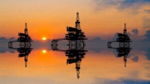 Latest U.S. Financial News and Analysis 25 Three offshore oil drilling platforms silhouetted against an orange sunset over calm water.