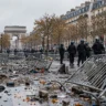 French police officers stand near overturned barricades and debris on a Paris street, with the Arc de Triomphe in the background.