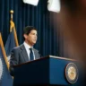 Federal Reserve Chair Kevin Warsh speaking at a podium with Federal Reserve flags behind him.