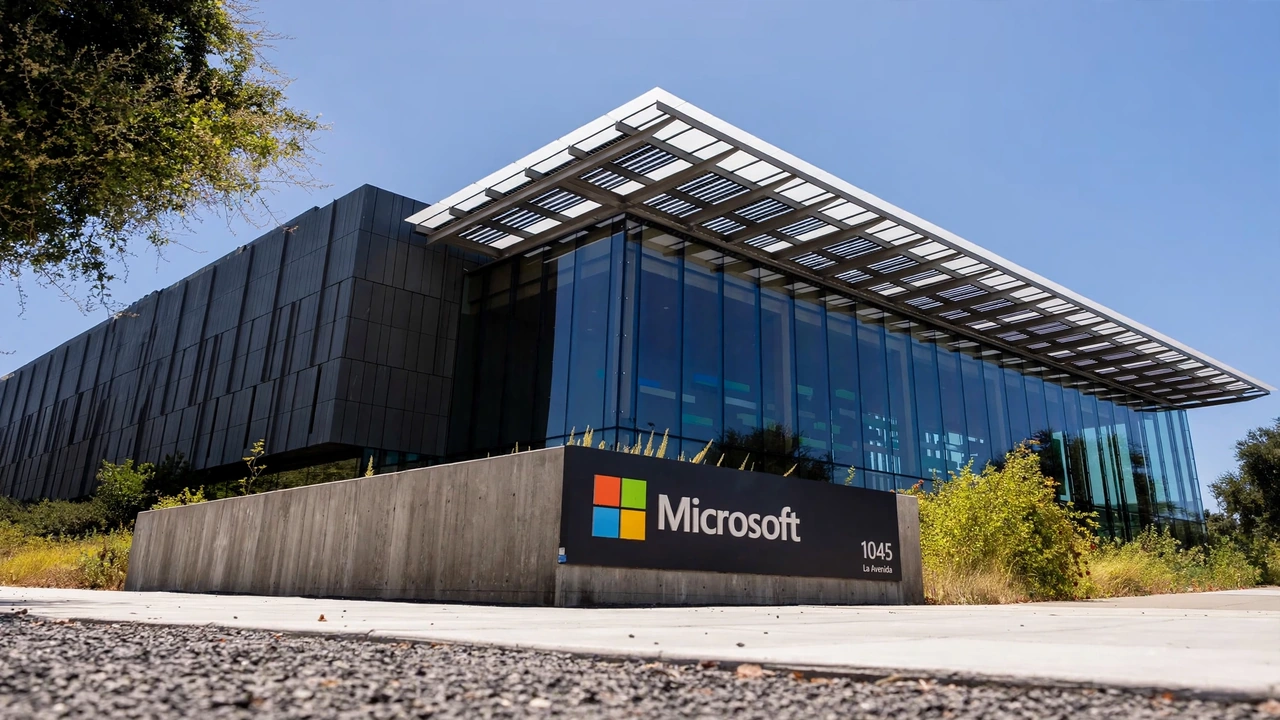Microsoft logo on a modern glass office building surrounded by landscaping under a clear blue sky.