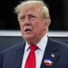 Donald Trump wearing an I Voted sticker and red tie with an American flag behind him.