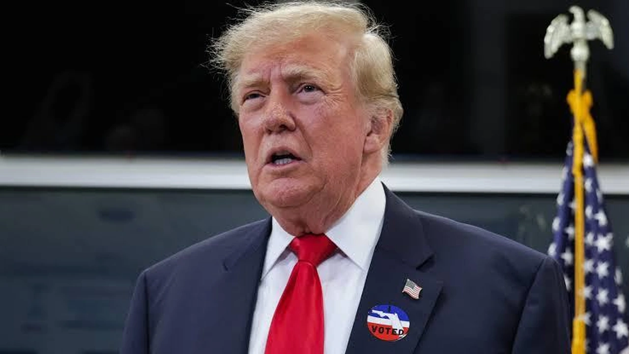 Donald Trump wearing an I Voted sticker and red tie with an American flag behind him.