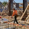 Construction worker wearing an orange safety vest and hard hat walks through an active building site with wooden supports and steel reinforcement.